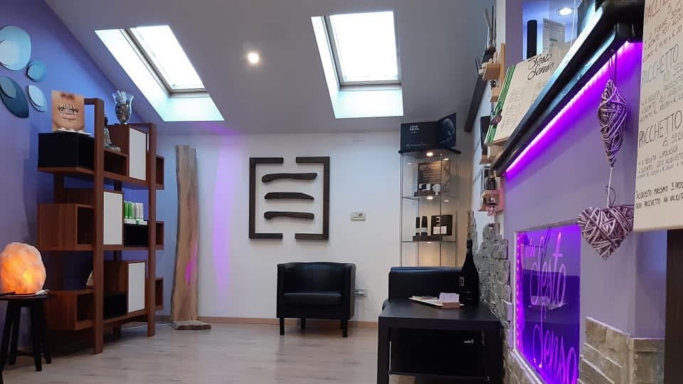La sala d'attesa di Estetica Sesto Senso: la poltrona e il tavolino sotto i lucernari, la scaffalatura dei prodotti e l'insegna «Sesto Senso» accesa sulla parete in pietra.
